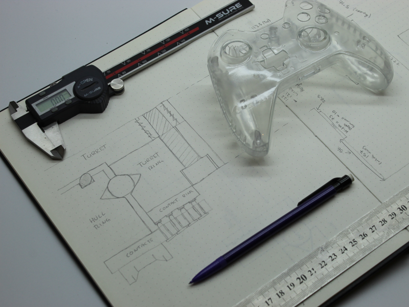 A engineers notepad with a 3D printed game controller with calipers and a pencil and ruler placed on top